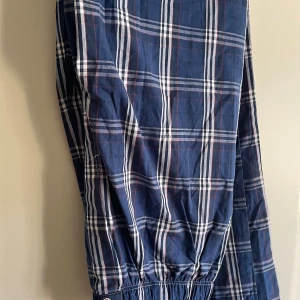 Gant tunna rutiga bomullsbyxor i Onesize - Snygga beg. blå rutiga byxor från Gant med elastisk midja och benavslut. Byxorna har ett klassiskt rutmönster i blått, vitt och rött och är tillverkade i mjuk bomull. Perfekta för en chill och avslappnad stil. Skicket är bra också, knappt använda.