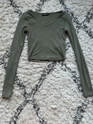 Olivgrön ribbad croppad topp FB Sister - Säljer en olivgrön croppad topp från FB Sister i storlek XS. Toppen är ribbad med v-ringning och har långa ärmar. Perfekt för dig som gillar en slim passform och enkel stil.