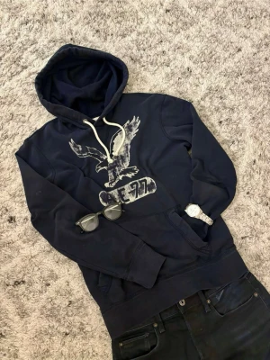 Mörkblå hoodie från American Eagle - Mörkblå hoodie med vit dragsko och stor örntryck på bröstet samt texten AE 77 | Size S | Otroligt limiterad hoodie, ställ frågor vid minsta lilla fundering!