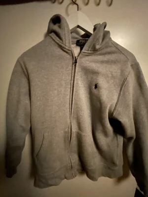 Grå hoodie från Polo Ralph Lauren - Säljer en grå hoodie från Polo Ralph Lauren med dragkedja och klassisk huva. Tröjan har två fickor fram och den ikoniska broderade loggan på bröstet. Materialet är mjukt och bekvämt, perfekt för chill dagar.