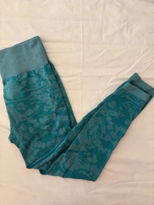 Turkosa leggings med mönster - Säljer ett par turkosa leggings med diskret mönster i ljusare blå toner. De har bred midjeresår för extra komfort och är tillverkade i ett stretchigt material som sitter snyggt. Perfekta för dig som vill ha något unikt och bekvämt.