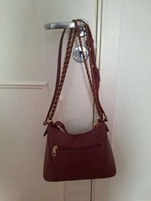 Vinröd axelväska  - Snygg vinröd axelväska i konstläder med guldfärgade kedjeremmar och dragkedja. Perfekt för dig som vill ha en praktisk väska med plats för det viktigaste.