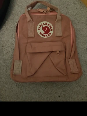 Rosa Kånken ryggsäck från Fjällräven - Säljer en klassisk rosa Kånken ryggsäck från Fjällräven med ikonisk rund logotyp framtill. Ryggsäcken har ett stort huvudfack, en mindre frontficka och slitstarka handtag och axelband i matchande färg. Materialet är tålig syntetväv.