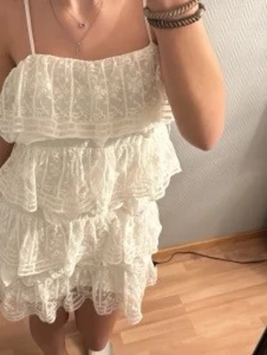Zara Vit volangklänning med spets - Söt vit kortklänning med tunna axelband och flera lager volanger. Klänningen har spetsdetaljer och är perfekt för sommarens varma dagar. Den är ärmlös och har en luftig passform som ger en romantisk känsla.