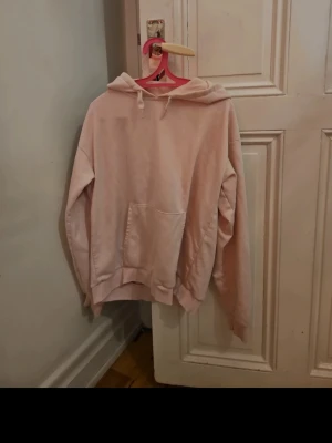 Ljusrosa hoodie med ficka - En ljusrosa hoodie med klassisk känguruficka framtill och justerbar huva med snören. Hoodien har ribbade muddar vid ärmslut och nederkant. Perfekt för en avslappnad och trendig look. Materialet känns mjukt och bekvämt, troligen bomullsblandning.