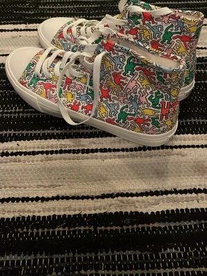 Färgglada high top sneakers - Snygga high top sneakers med färgglatt Keith Haring-inspirerat mönster i rött, gult, grönt och blått på vit bas. Skorna har vita snören och sula, samt klassisk rund tå. Perfekta för dig som vill sticka ut med din stil.