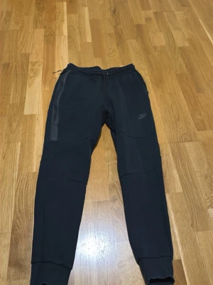 Svarta Nike mjukisbyxor med logga - Svarta mjukisbyxor från Nike med diskret logga på vänster ben. Byxorna har dragsko i midjan, ribbade muddar vid benslut och praktiska sidofickor. Materialet är mjukt och bekvämt, perfekt för chill eller träning.