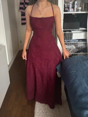 Vinröd långklänning med snörning - Vinröd långklänning med tunna axelband och öppen rygg med snygg snörning. Klänningen har en elegant, figurnära passform upptill och faller fint nedtill. Perfekt för speciella tillfällen.
