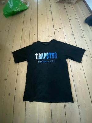 Svart Trapstar t-shirt - Cool svart t-shirt från Trapstar med stort tryck i vitt och blått på bröstet där det står 'Trapstar' och 'It's a secret'. T-shirten har korta ärmar och en klassisk passform. Perfekt för dig som gillar streetwear och vill sticka ut.