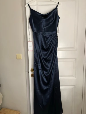 Marinblå långklänning från JJ’s house - Elegant marinblå långklänning från JJ’s house med smala axelband och draperad detalj framtill. Klänningen har en slät, glansig yta som ger en lyxig känsla och är tillverkad i satinliknande material. Perfekt för dig som vill ha en stilren och modern look.
