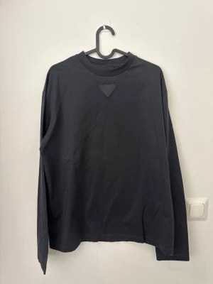 Svart långärmad t-shirt från Prada - Snygg svart långärmad t-shirt från Prada med en ikonisk trekantig patch på bröstet. Tillverkad i mjuk bomull och har en klassisk rund halsringning. Perfekt för dig som gillar stilrena och exklusiva plagg.