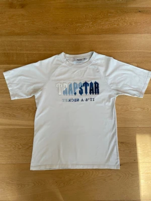 Vit Trapstar t-shirt med blått tryck - Vit t-shirt från Trapstar med korta ärmar och tryck i blått och vitt framtill. Klassisk passform och rund halsringning. Perfekt för en avslappnad streetwear-look.
