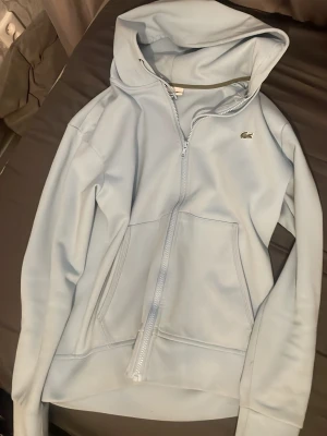 Ljusblå hoodie från Lacoste - Snygg ljusblå hoodie från Lacoste med dragkedja framtill och klassisk krokodillogga på bröstet. Hoodien har två stora fickor och en skön huva med dragsko. Perfekt för en chill och stilren look.