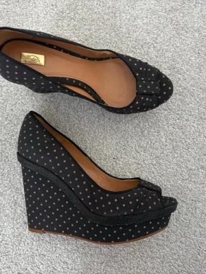 Svarta pumps med kilklack och prickar - Säljer ett par svarta pumps från Salvatore Ferragamo med hög kilklack och peep-toe. Skorna är täckta av ett tyg med ljusrosa prickar och har läder på insidan. Perfekt för dig som gillar en lekfull och retro look. Finns tydliga tecken på användning. Köpt i Spanien.