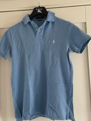 Blå pike från Polo Ralph Lauren - Snygg ljusblå pike från Polo Ralph Lauren med klassisk krage och två knappar framtill. T-shirten har korta ärmar och ikonisk logga broderad på bröstet. Perfekt för dig som gillar stilrena och tidlösa plagg.