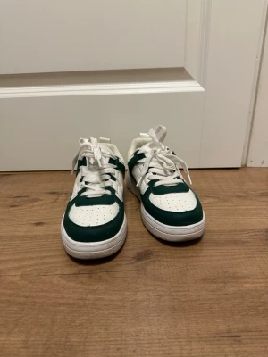 Vita och gröna sneakers - Snygga sneakers i vitt och grönt med klassisk låg siluett. Skorna har perforerad tå, vita skosnören och paneler i konstläder. Perfekta för dig som gillar streetwear och vill sticka ut lite extra.