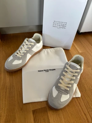 Maison Margiela vita och grå sneakers  - Maison Margiela sneakers i vitt och ljusgrått med klassisk låg silhuett. Ovandel i mocka och läder, vita skosnören och brun gummisula. Snygg och stilren design som passar till det mesta. Kommer med dustbag och originalkartong.