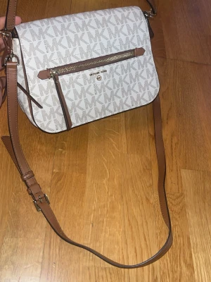Michael Kors axelväska beige/brun (ny skick) - Snygg axelväska från Michael Kors i beige med MK-logga över hela väskan. Väskan har bruna detaljer, justerbar axelrem och en praktisk ficka med dragkedja framtill. Tillverkad i tåligt konstläder och har guldfärgade metalldetaljer. 100% äkta, ny skick.