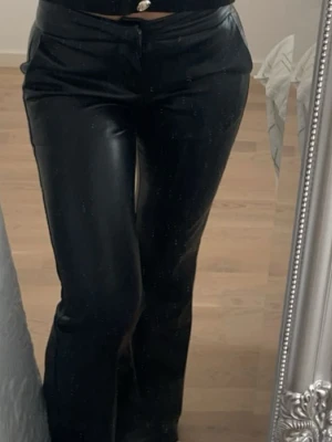 Lågmidjade utsvängda skinn byxor - Säljer ett par svarta coated jeans med rak passform och hög midja. Jeansen har en glansig finish som ger en cool look och är perfekta för att lyfta vilken outfit som helst. Materialet är troligtvis en blandning av bomull och polyester med en coated yta. Storlek Xs, passar i mig som är 1,64 cm, från bershka