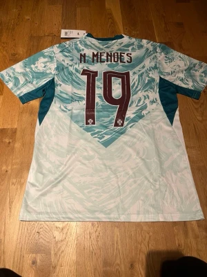 Portugal bortatröja Puma N Mendes #19 - Säljer en Portugal bortatröja från Puma med N Mendes och nummer 19 på ryggen. Tröjan har ett unikt turkost och vitt mönster, korta ärmar och Portugal-emblemet på bröstet. Tillverkad i lätt och ventilerande material, perfekt för fotboll eller träning.