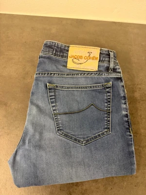 Blå jeans från Jacob Cohen W34 - Snygga blå jeans från Jacob Cohen med klassisk femficksmodell och raka ben. Jeansen har en ljus tvätt och subtila slitningar för en avslappnad look. Tillverkade i mjukt denimtyg med patch i mockaimitation bak på linningen.