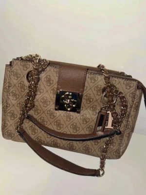 Beige och brun handväska från Guess - Säljer fin Guess väska som rymmer ganska mycket. Använd men i skick som ny.  Modellen: Guess logo city sml society satchel   Ny pris: 1500kr 