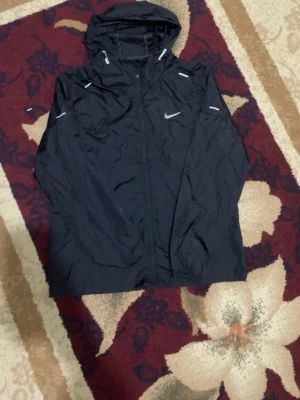 Svart regnjacka från Nike med huva - Snygg svart regnjacka från Nike med dragkedja framtill och justerbar huva. Jackan har vita detaljer och Nike-logga på bröstet. Tillverkad i lätt, vattenavvisande material som passar perfekt för regniga dagar.