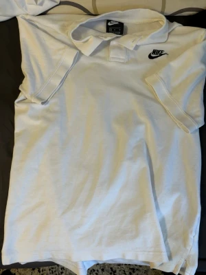 Vit Nike pikétröja med logga - Säljer en klassisk vit pikétröja från Nike med svart broderad logga på bröstet. Tröjan har korta ärmar, krage och knappar framtill. Perfekt för dig som gillar sportig och stilren look.