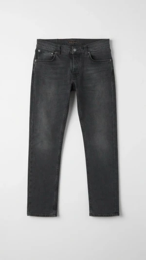 Grå Nudie Jeans Grim Tim - Säljer ett par svarta slim fit jeans från Nudie Jeans i modellen Grim Tim med klassisk femficksdesign och knappgylf. Jeansen har en lätt tvättad look och är tillverkade i bomull med stretch för extra komfort. Perfekt för dig som gillar en stilren och modern look.