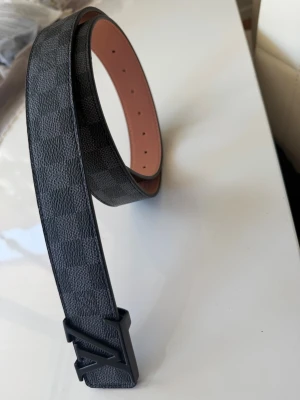 Louis Vuitton Damier svart bälte - Snyggt svart bälte från Louis Vuitton med klassiskt Damier-mönster och en stilren LV-logga i svart metall som spänne. Bältet är tillverkat i läder och har en slät, matt finish på insidan. Perfekt för att lyfta din outfit med en exklusiv touch.Pris kan diskuteras 😁