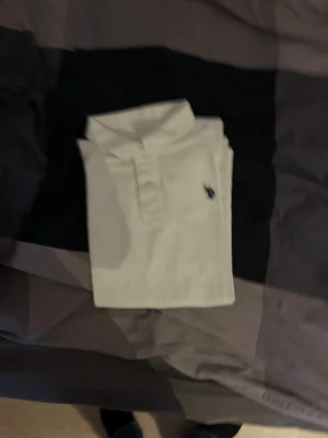 Vit polotopp från us polo assn  - Säljer en klassisk vit polotopp från us polo assn med krage och knappar framtill. Toppens ikoniska logga är broderad på bröstet. Perfekt för dig som gillar stilrena och tidlösa plagg. Materialet är mjukt och känns bekvämt mot huden.