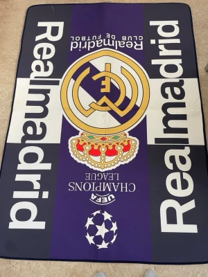 Real Madrid filt med UEFA Champions League - Stor filt med Real Madrid-logga och UEFA Champions League-motiv. Färgerna är svart, vit, lila och guld med tydliga tryck och klubbnamnet på sidorna. Perfekt för fotbollsfans som vill visa sitt stöd för laget.