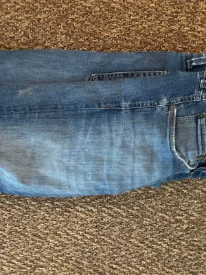 Blå jeans från Jack and Jones  - Säljer ett par klassiska blå jeans från Jack and Jones med snyggt slitna detaljer och femficksdesign. Jeansen har en ljusblå tvätt och är tillverkade i bomull. Bakfickan har Jeans karakteristiska broderi. Perfekt för dig som gillar en avslappnad stil.
