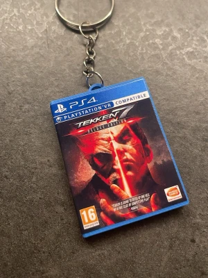 Tekken 7 nyckelring PS4-fodral - Unik nyckelring formad som ett PS4-fodral för Tekken 7 Deluxe Edition. Fodralet är i miniatyr och har detaljerad framsida och baksida med färgstarkt tryck. Perfekt accessoar för gamers som vill visa sin kärlek till fighting-spel. Metallkedja och ring för enkel fastsättning.