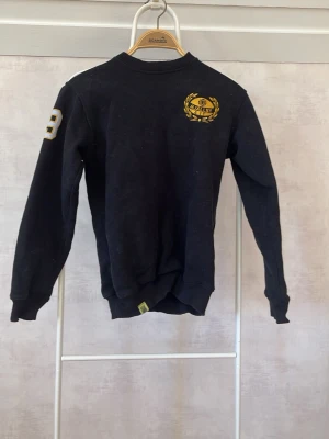 Svart sweatshirt med tryck från Mjällby AIF - Cool svart sweatshirt från Mjällby AIF med gult tryck och logga på bröstet samt siffran 39 på ena ärmen. Tröjan har rund hals, långa ärmar och ribbade muddar. Perfekt för en avslappnad och sportig stil.
