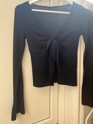 Svart knytblus från Gina Tricot - Trendig svart långärmad topp från Gina Tricot med snygg knytning framtill och djup v-ringning. Tillverkad i mjukt material som sitter skönt på kroppen. Perfekt för dig som gillar stilrena och enkla plagg med en twist.