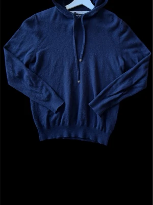 Mörkblå hoodie i mjuk ullmix - Snygg mörkblå hoodie med huva och dragsko. Tillverkad i en mjuk och skön ullmix som känns lyxig mot huden. Långa ärmar och ribbade muddar vid ärmslut och nederkant ger en clean look. Perfekt för dig som gillar stilrena och bekväma plagg.