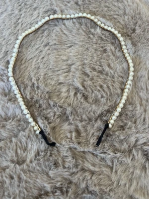 Vågigt diadem med pärlor - Ett snyggt diadem med vågig form, dekorerat med vita pärlor längs hela bågen. Metallstommen är guldfärgad och diademet har svarta detaljer vid ändarna. Perfekt för att ge en elegant touch till din look.