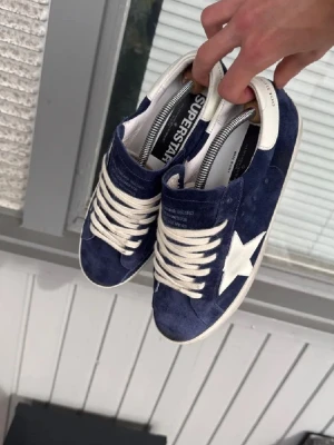 Blåa Golden Goose Superstar sneakers - Säljer ett par blåa Golden Goose Superstar sneakers i mocka med vit stjärndetalj på sidan, vita skosnören och vit sula. Skorna har lågt skaft och klassisk retrostil. Perfekta för dig som gillar streetwear och vill ha något unikt.