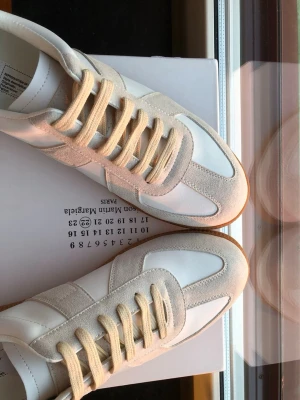 Maison Margiela vita sneakers - Maison Margiela sneakers i vitt och beige med klassisk design. Skorna har en kombination av slätt läder och mocka, beige snörning och gummisula. Perfekt för dig som gillar stilrena och trendiga sneakers med premiumkänsla.