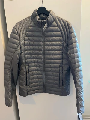 Grå dunjacka från Colmar - Säljer en grå dunjacka från Colmar med quiltad design och hög krage. Jackan har två sidofickor med dragkedja och Colmar-logga på vänster ärm. Storlek 48