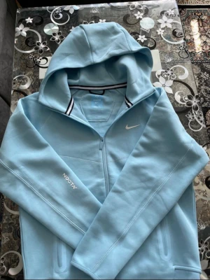 Nike NOCTA ljusblå hoodie - Ljusblå hoodie från Nike NOCTA med dragkedja och huva. Diskret Nike-logga på bröstet och NOCTA-tryck på ärmen. Mjukt material och sportig design med svarta och vita detaljer i kragen. Perfekt för dig som gillar streetwear och exklusiva samarbeten.