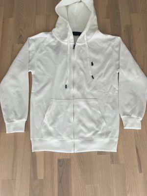 Vit hoodie från Polo Ralph Lauren - Vit hoodie från Polo Ralph Lauren med dragkedja, huva med snören och klassisk logga broderad på bröstet. Tillverkad i mjukt material och har en stor ficka framtill. Perfekt för en chill och stilren look.
