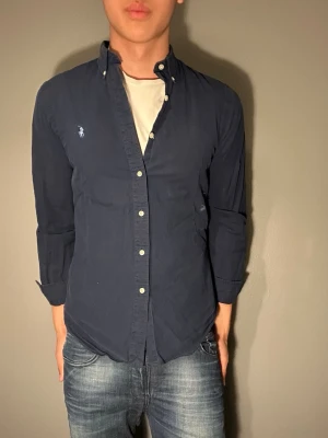 Marinblå skjorta från Polo Ralph Lauren - Säljer en klassisk marinblå skjorta från Polo Ralph Lauren med vit broderad logga på bröstet. Skjortan har knappar framtill och långärmad design, perfekt för dig som gillar stilrena plagg med ikonisk branding.