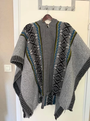 Grå mönstrad sjal poncho med huva - Säljer en stor och mysig grå sjal med huva och dekorativa mönster i svart, blått och grönt. Sjalen har ett klassiskt geometriskt mönster och är tillverkad i ett stickat material som känns både varmt och bekvämt. Perfekt att slänga över axlarna för en cool look.