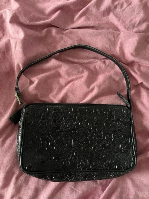Svart väska med spetsmönster - Elegant svart kuvertväska från La Bagagerie med präglat mönster och justerbar axelrem. 