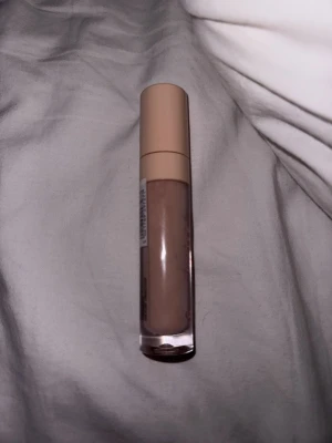 Beige concealer med nude lock - Säljer en flytande concealer i beige nyans med nude-färgat lock. Förpackningen är cylinderformad och genomskinlig så man ser färgen på produkten. Perfekt för att täcka mörka ringar eller blemmor. Enkel att applicera med applikator.