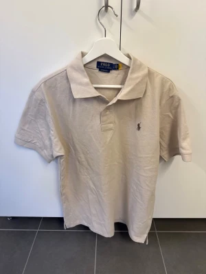 Beige piké från Polo Ralph Lauren - Snygg beige pikétröja från Polo Ralph Lauren i custom slim fit. Klassisk krage, korta ärmar och den ikoniska broderade loggan på bröstet. Perfekt till jeans eller shorts för en clean och stilren look.