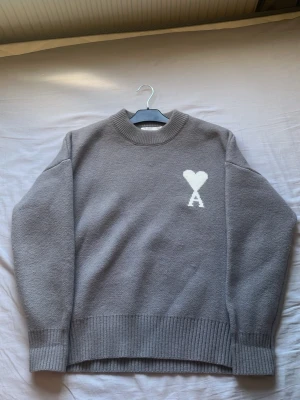 Grå stickad tröja från Ami Paris - Säljer en grå stickad tröja från Ami Paris med ett vitt hjärta och bokstaven A broderat på bröstet. Tröjan har rund hals, ribbade muddar och är tillverkad i mjukt material. Perfekt för dig som gillar stilrena plagg med diskret detalj.