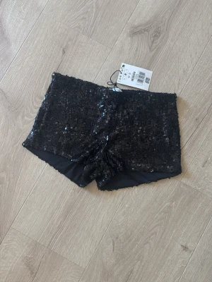 Svarta paljettshorts från Gina Tricot - Säljer ett par svarta shorts från Gina Tricot med glittriga paljetter över hela ytan. De har en tight passform. Shortsen har en hög midja och är tillverkade i polyester för en bekväm känsla.